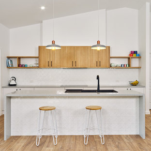 This is an example of a mid-sized contemporary galley open plan kitchen in Other with a drop-in sink, flat-panel cabinets, light wood cabinets, quartz benchtops, white splashback, black appliances, laminate floors, with island, brown floor and grey benchtop.