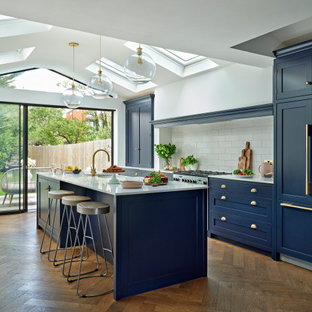 Photo of a large traditional galley kitchen/diner in London with a belfast sink, shaker cabinets, blue cabinets, quartz worktops, white splashback, ceramic splashback, integrated appliances, an island, white worktops, a vaulted ceiling, medium hardwood flooring and brown floors.