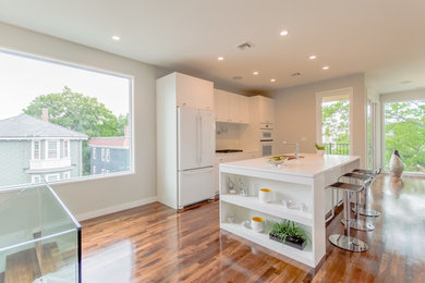 Example of a large trendy l-shaped dark wood floor open concept kitchen design in Boston with an integrated sink, flat-panel cabinets, white cabinets, quartz countertops, white backsplash, glass tile backsplash, white appliances and an island