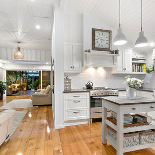 Photo of a small beach style u-shaped open plan kitchen in Brisbane with a farmhouse sink, white cabinets, granite benchtops, white splashback, ceramic splashback, stainless steel appliances, with island, recessed-panel cabinets, medium hardwood floors, brown floor and grey benchtop.