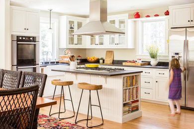 Exemple d'une cuisine américaine chic en L de taille moyenne avec un évier de ferme, un placard à porte vitrée, des portes de placard blanches, un électroménager en acier inoxydable, parquet clair, îlot, un plan de travail en granite, fenêtre et un sol marron.