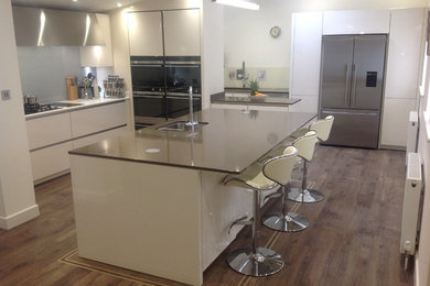 Large contemporary u-shaped kitchen/diner in Surrey with a double-bowl sink, flat-panel cabinets, white cabinets, composite countertops, metallic splashback, glass sheet splashback, black appliances and an island.
