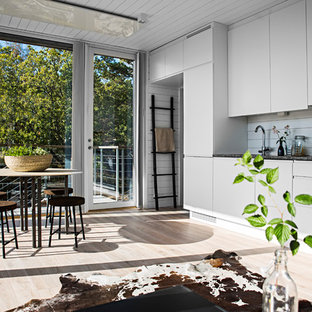 Inspiration for a mid-sized scandinavian galley open plan kitchen in Stockholm with a drop-in sink, flat-panel cabinets, white cabinets, limestone benchtops, white splashback, glass sheet splashback, white appliances and light hardwood floors.