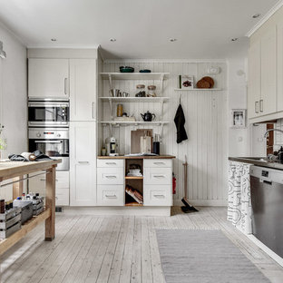 This is an example of a mid-sized country l-shaped separate kitchen in Stockholm with shaker cabinets, beige cabinets, wood benchtops, white splashback, stainless steel appliances, light hardwood floors and with island.