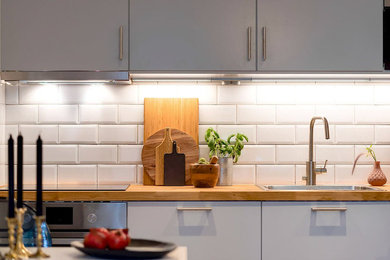 Bild på ett litet funkis linjärt kök med öppen planlösning, med en enkel diskho, släta luckor, grå skåp, träbänkskiva, vitt stänkskydd, stänkskydd i keramik, rostfria vitvaror och mellanmörkt trägolv