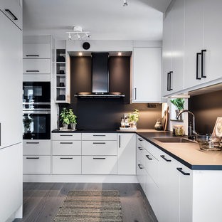 Inredning av ett minimalistiskt stort kök, med en integrerad diskho, släta luckor, vita skåp, svart stänkskydd, svarta vitvaror, grått golv och ljust trägolv