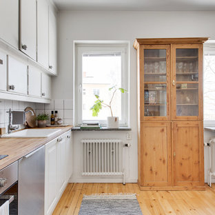 Bild på ett minimalistiskt beige linjärt beige kök, med en nedsänkt diskho, luckor med glaspanel, vita skåp, träbänkskiva, vitt stänkskydd, stänkskydd i keramik, rostfria vitvaror, ljust trägolv och beiget golv