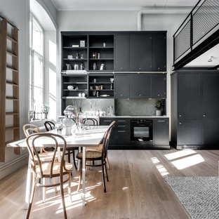 Photo of a mid-sized scandinavian single-wall open plan kitchen in Stockholm with limestone benchtops, a farmhouse sink, flat-panel cabinets, black cabinets, grey splashback, stone slab splashback, black appliances, light hardwood floors and no island.