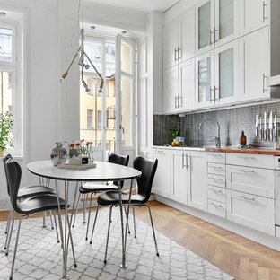 Skandinavisk inredning av ett stort linjärt kök och matrum, med vita skåp, träbänkskiva, stänkskydd i mosaik, vita vitvaror, svart stänkskydd, ljust trägolv och skåp i shakerstil