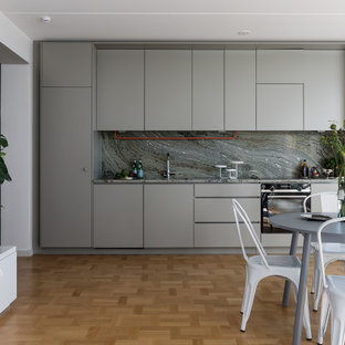 Minimalistisk inredning av ett grå linjärt grått kök med öppen planlösning, med släta luckor, grå skåp, grått stänkskydd, stänkskydd i sten, rostfria vitvaror, mellanmörkt trägolv och brunt golv