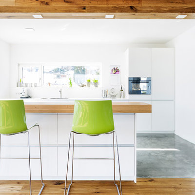 Inspiration for a mid-sized contemporary galley concrete floor and gray floor open concept kitchen remodel in Stuttgart with a single-bowl sink, flat-panel cabinets, white cabinets, stainless steel countertops, white backsplash, glass sheet backsplash, black appliances, a peninsula and white countertops