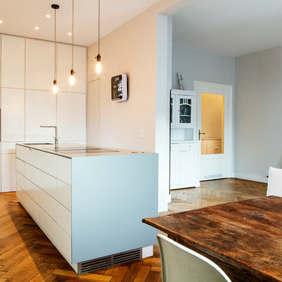 Example of a mid-sized trendy single-wall medium tone wood floor and brown floor open concept kitchen design in Munich with flat-panel cabinets, white cabinets, an island, an integrated sink, solid surface countertops, white backsplash, limestone backsplash and stainless steel appliances