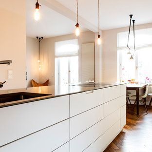 Modelo de cocina lineal, contemporánea, de tamaño medio, abierta, con fregadero integrado, armarios con paneles lisos, puertas de armario blancas, encimera de acrílico, salpicadero blanco, salpicadero de piedra caliza, electrodomésticos de acero inoxidable, suelo de madera en tonos medios, una isla y suelo marrón