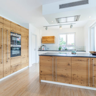 Große Moderne Küche in L-Form mit flächenbündigen Schrankfronten, hellbraunen Holzschränken, Küchenrückwand in Weiß, Glasrückwand, schwarzen Elektrogeräten, hellem Holzboden, Halbinsel, beigem Boden und schwarzer Arbeitsplatte in Sonstige