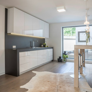 Offene, Einzeilige, Kleine Moderne Küche ohne Insel mit Einbauwaschbecken, flächenbündigen Schrankfronten, weißen Schränken, Küchenrückwand in Grau, braunem Holzboden und braunem Boden in Essen