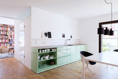 Mittelgroße Moderne Wohnküche ohne Insel mit integriertem Waschbecken, Edelstahl-Arbeitsplatte, Küchenrückwand in Weiß, Elektrogeräten mit Frontblende und hellem Holzboden in München