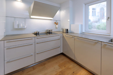 This is an example of a medium sized contemporary u-shaped kitchen in Bremen with no island, white splashback, black appliances, a built-in sink, flat-panel cabinets, white cabinets, light hardwood flooring and brown floors.