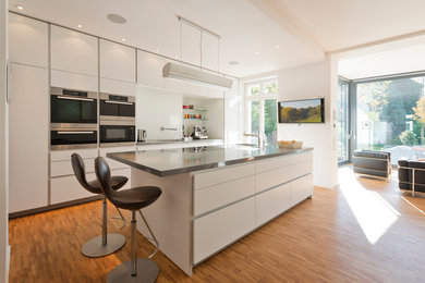 Large contemporary open plan kitchen in Berlin with a submerged sink, flat-panel cabinets, white cabinets, white splashback, glass sheet splashback, stainless steel appliances, medium hardwood flooring and an island.