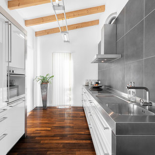 Zweizeilige Moderne Küche ohne Insel mit Edelstahl-Arbeitsplatte, Küchenrückwand in Grau, Rückwand aus Keramikfliesen, dunklem Holzboden, braunem Boden, grauer Arbeitsplatte, integriertem Waschbecken, flächenbündigen Schrankfronten und weißen Schränken in München