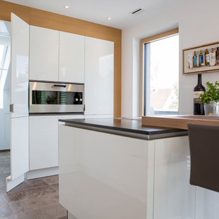 This is an example of a mid-sized contemporary l-shaped open plan kitchen in Frankfurt with a drop-in sink, flat-panel cabinets, white cabinets, granite benchtops, black splashback, glass sheet splashback, stainless steel appliances, terra-cotta floors, a peninsula, brown floor and black benchtop.
