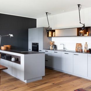 Zweizeilige, Mittelgroße Moderne Wohnküche mit Waschbecken, flächenbündigen Schrankfronten, grauen Schränken, Küchenrückwand in Weiß, schwarzen Elektrogeräten, hellem Holzboden, Halbinsel und schwarzer Arbeitsplatte in Köln