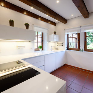 Mittelgroße Landhausstil Küche in U-Form mit integriertem Waschbecken, flächenbündigen Schrankfronten, weißen Schränken, Küchenrückwand in Weiß, Glasrückwand, schwarzen Elektrogeräten, Terrakottaboden, Halbinsel, rotem Boden und weißer Arbeitsplatte in Essen