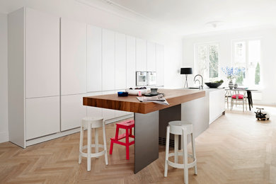 Modelo de cocina comedor moderna grande con armarios con paneles lisos, puertas de armario blancas, suelo de madera clara, una isla, fregadero bajoencimera y electrodomésticos con paneles