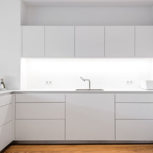 Offene, Mittelgroße Moderne Küche in L-Form mit Waschbecken, flächenbündigen Schrankfronten, weißen Schränken, Mineralwerkstoff-Arbeitsplatte, Küchenrückwand in Weiß, Glasrückwand, weißen Elektrogeräten, hellem Holzboden, Halbinsel und weißer Arbeitsplatte in Hamburg