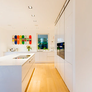 Offene, Zweizeilige, Große Moderne Küche mit Einbauwaschbecken, flächenbündigen Schrankfronten, weißen Schränken, Mineralwerkstoff-Arbeitsplatte, Küchenrückwand in Weiß, Glasrückwand, schwarzen Elektrogeräten, hellem Holzboden, Kücheninsel, beigem Boden und weißer Arbeitsplatte in Düsseldorf