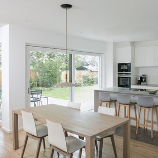 Mittelgroße, Einzeilige Moderne Wohnküche mit Unterbauwaschbecken, flächenbündigen Schrankfronten, weißen Schränken, Küchenrückwand in Weiß, Glasrückwand, schwarzen Elektrogeräten, hellem Holzboden, Kücheninsel, beigem Boden, grauer Arbeitsplatte und Mineralwerkstoff-Arbeitsplatte in Düsseldorf