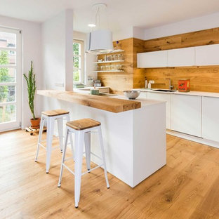 Mittelgroße Nordische Küche mit flächenbündigen Schrankfronten, weißen Schränken, Rückwand aus Holz, braunem Holzboden, Halbinsel, weißer Arbeitsplatte und Unterbauwaschbecken in Nürnberg