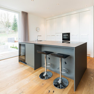 Offene, Mittelgroße, Zweizeilige Moderne Küche mit Unterbauwaschbecken, flächenbündigen Schrankfronten, weißen Schränken, Laminat-Arbeitsplatte, schwarzen Elektrogeräten, braunem Holzboden, Kücheninsel, braunem Boden und grauer Arbeitsplatte in München