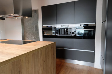 This is an example of a medium sized contemporary l-shaped enclosed kitchen in Other with flat-panel cabinets, black cabinets, wood worktops, an island, beige worktops, stainless steel appliances, a built-in sink, medium hardwood flooring and beige floors.