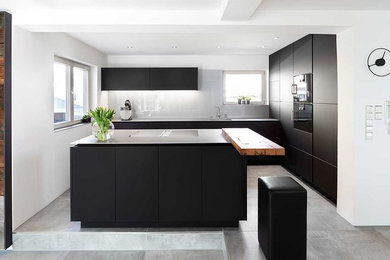 Foto på ett mellanstort funkis kök, med släta luckor, svarta skåp, bänkskiva i rostfritt stål, glaspanel som stänkskydd, svarta vitvaror, betonggolv, en köksö, grått golv och vitt stänkskydd