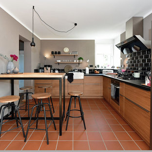 Inspiration for a mid-sized industrial l-shaped open plan kitchen in Frankfurt with a single-bowl sink, flat-panel cabinets, medium wood cabinets, wood benchtops, black splashback, subway tile splashback, stainless steel appliances, terra-cotta floors, a peninsula, red floor and black benchtop.