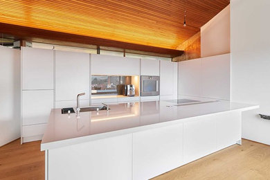 Offene, Einzeilige Moderne Küche mit flächenbündigen Schrankfronten, weißen Schränken, braunem Holzboden, Kücheninsel, braunem Boden, Einbauwaschbecken und Küchengeräten aus Edelstahl in Stuttgart