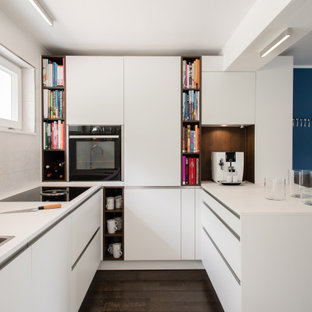 Moderne Küche in U-Form mit Einbauwaschbecken, flächenbündigen Schrankfronten, weißen Schränken, schwarzen Elektrogeräten, dunklem Holzboden, Halbinsel, braunem Boden und weißer Arbeitsplatte in Stuttgart