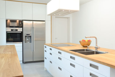 Geschlossene Moderne Küche mit Doppelwaschbecken, weißen Schränken, Arbeitsplatte aus Holz, Küchenrückwand in Weiß, Küchengeräten aus Edelstahl, Betonboden, Kücheninsel, grauem Boden und brauner Arbeitsplatte in München
