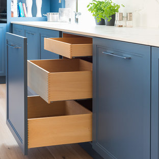 Design ideas for a mid-sized transitional kitchen in Munich with an integrated sink, shaker cabinets, blue cabinets, white splashback, black appliances, medium hardwood floors, with island, brown floor and white benchtop.