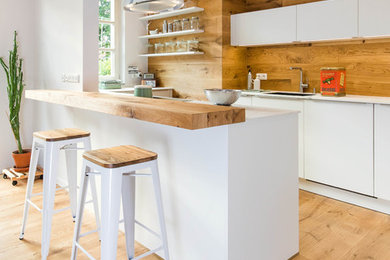 Bild på ett mellanstort skandinaviskt vit vitt kök, med en integrerad diskho, släta luckor, vita skåp, brunt stänkskydd, stänkskydd i trä, mellanmörkt trägolv, en halv köksö och brunt golv
