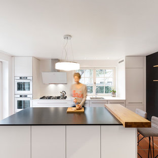 Offene, Zweizeilige, Mittelgroße Moderne Küche mit Waschbecken, flächenbündigen Schrankfronten, weißen Schränken, Granit-Arbeitsplatte, Küchenrückwand in Weiß, Rückwand aus Holz, weißen Elektrogeräten, Kücheninsel, schwarzer Arbeitsplatte und rotem Boden in Stuttgart