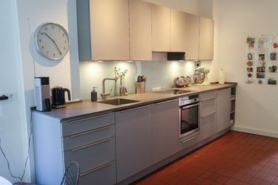 ハンブルクにあるお手頃価格の小さなコンテンポラリースタイルのおしゃれなキッチン (シングルシンク、フラットパネル扉のキャビネット、グレーのキャビネット、ガラス板のキッチンパネル、シルバーの調理設備、セラミックタイルの床、アイランドなし、赤い床、白いキッチンパネル) の写真