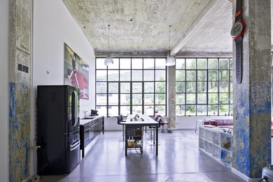 他の地域にある広いインダストリアルスタイルのおしゃれなキッチン (フラットパネル扉のキャビネット、黒いキャビネット、黒い調理設備、コンクリートの床、白いキッチンパネル) の写真