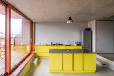 ベルリンにあるコンテンポラリースタイルのおしゃれなキッチン (シングルシンク、フラットパネル扉のキャビネット、黄色いキャビネット、ステンレスカウンター、白いキッチンパネル、シルバーの調理設備、コンクリートの床) の写真