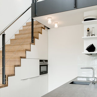 Zweizeilige, Kleine, Offene Industrial Küche mit flächenbündigen Schrankfronten, weißen Schränken, Betonarbeitsplatte, Küchenrückwand in Weiß, Glasrückwand, Halbinsel, integriertem Waschbecken und schwarzen Elektrogeräten in Hamburg