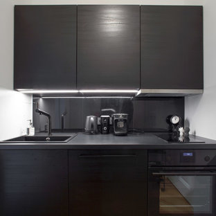 Modelo de cocina lineal, minimalista, pequeña, cerrada, sin isla, con fregadero integrado, armarios con puertas mallorquinas, puertas de armario negras, encimera de laminado, salpicadero negro, salpicadero de metal, electrodomésticos negros, suelo de madera en tonos medios y suelo marrón