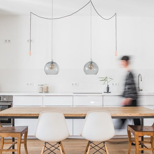 Einzeilige, Große Moderne Wohnküche ohne Insel mit flächenbündigen Schrankfronten, weißen Schränken, Mineralwerkstoff-Arbeitsplatte, Küchenrückwand in Weiß, Rückwand aus Porzellanfliesen, Küchengeräten aus Edelstahl, braunem Holzboden, braunem Boden, weißer Arbeitsplatte und Unterbauwaschbecken in Berlin