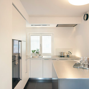 Ispirazione per una piccola cucina minimal chiusa con lavello integrato, ante lisce, ante bianche, top in acciaio inossidabile, paraspruzzi bianco, paraspruzzi con lastra di vetro, elettrodomestici neri, isola e pavimento in cemento