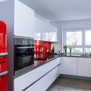 Mittelgroße Moderne Küche ohne Insel in U-Form mit integriertem Waschbecken, flächenbündigen Schrankfronten, weißen Schränken, Küchenrückwand in Rot, Glasrückwand, bunten Elektrogeräten und hellem Holzboden in München