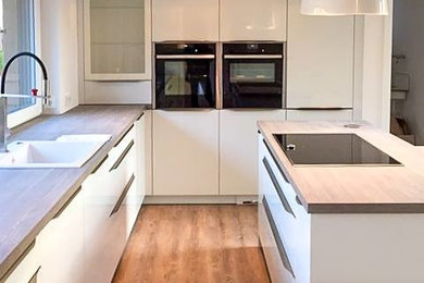 Design ideas for a medium sized contemporary l-shaped open plan kitchen in Hamburg with a built-in sink, flat-panel cabinets, white cabinets, wood worktops, white splashback, black appliances, light hardwood flooring, an island and brown floors.